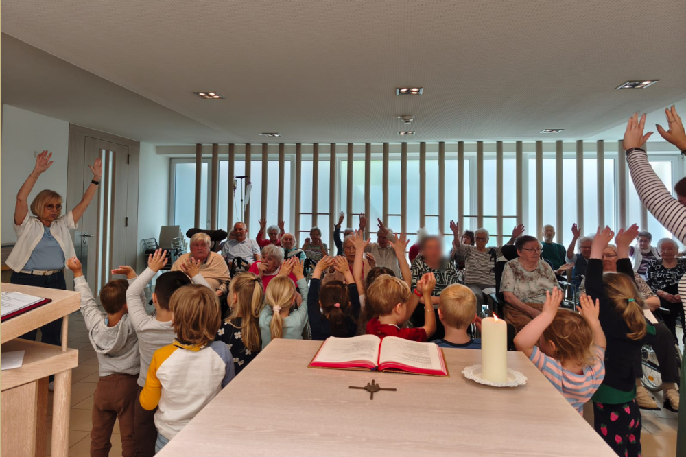 Eine bunte Gruppe aus Kindern und älteren Menschen versammelt sich in einem hellen, modernen Raum mit großen Fenstern und Holzvertäfelung.  Alle Teilnehmer heben die Hände. Im Vordergrund befindet sich ein mit einem offenen Buch, einer weißen Kerze auf einem Teller, einem holzfarbenen Kreuz.